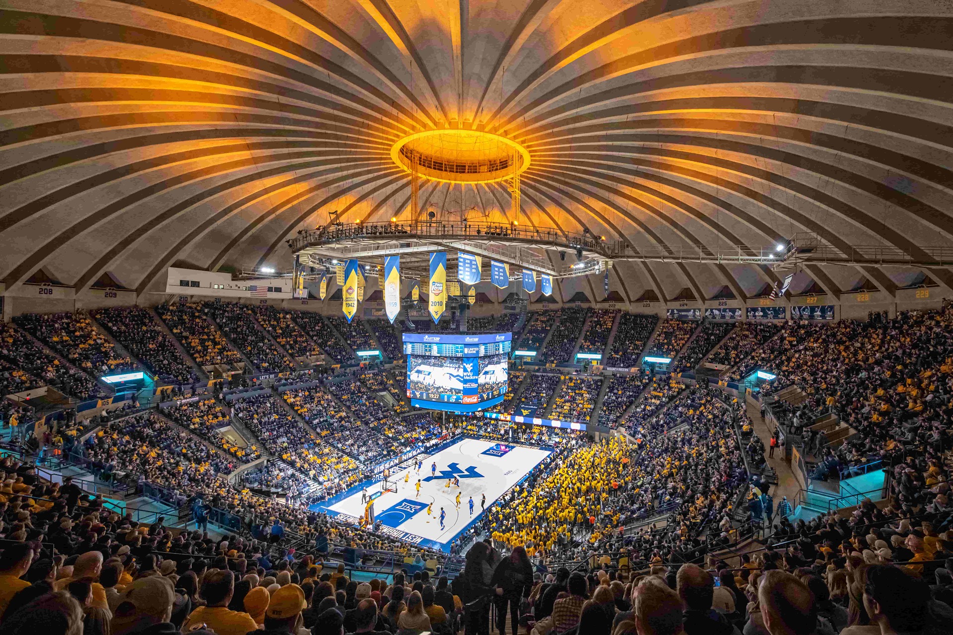 Wide interior view of the WVU Coliseum during a packed West Virginia University basketball game, with fans filling the stands around the court and the arena’s distinctive domed ceiling glowing gold above the center scoreboard.