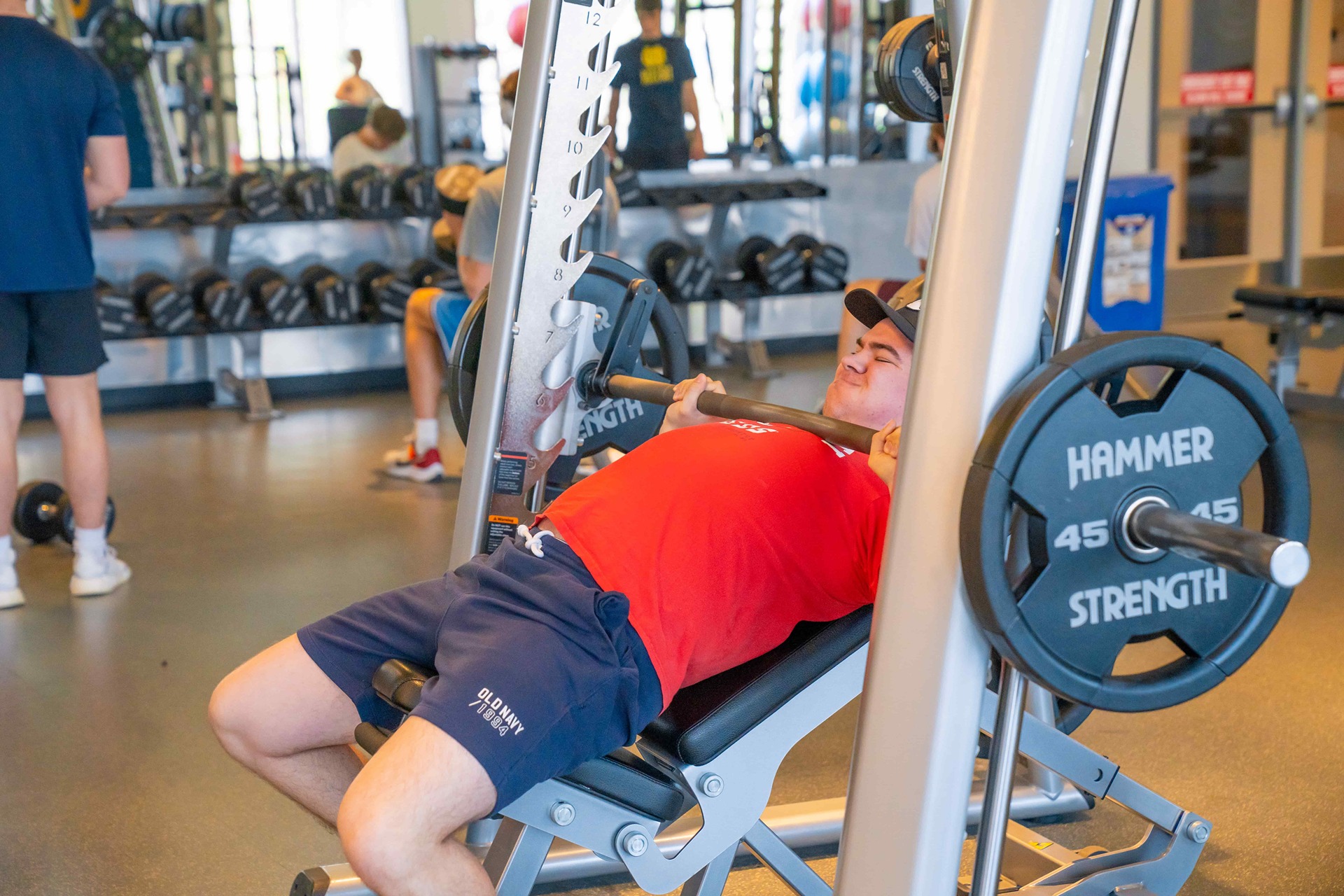 A WVU student performs a bench press on a Smith machine at the Downtown Fitness Center. He is lying on a slightly inclined bench, wearing a red T-shirt, navy shorts and a black cap, pressing a barbell loaded with 45-pound plates. Other students and free weights are visible in the background of the gym.