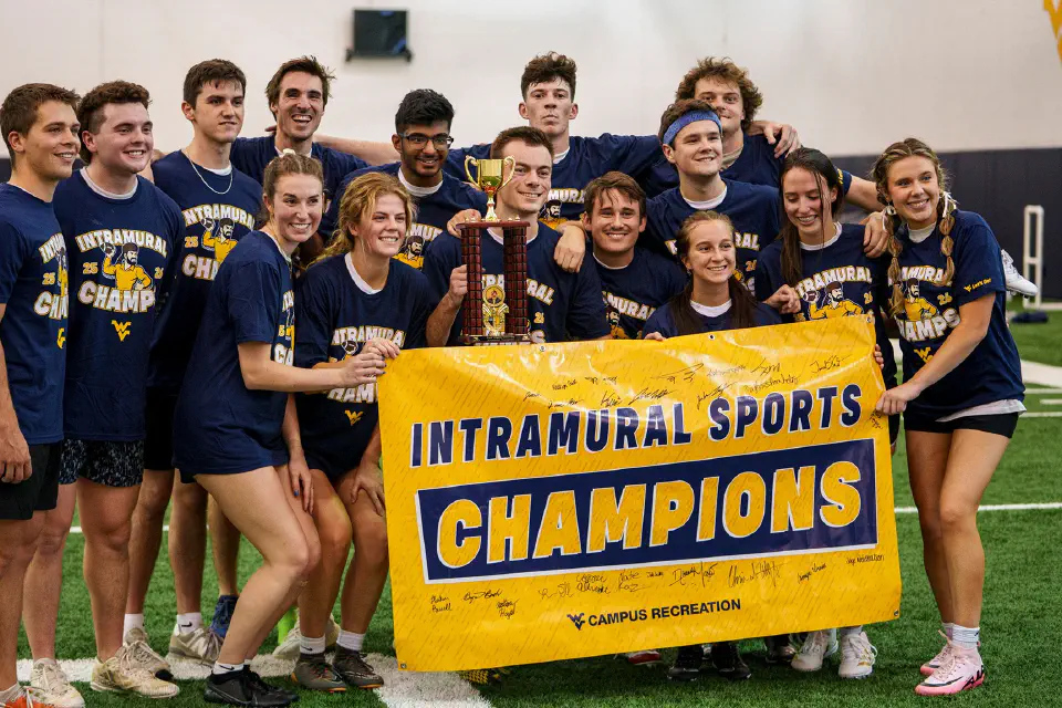 A group of college students wearing matching “Intramural Champs” shirts pose on an indoor turf field, smiling and holding a large “Intramural Sports Champions” banner and a trophy.