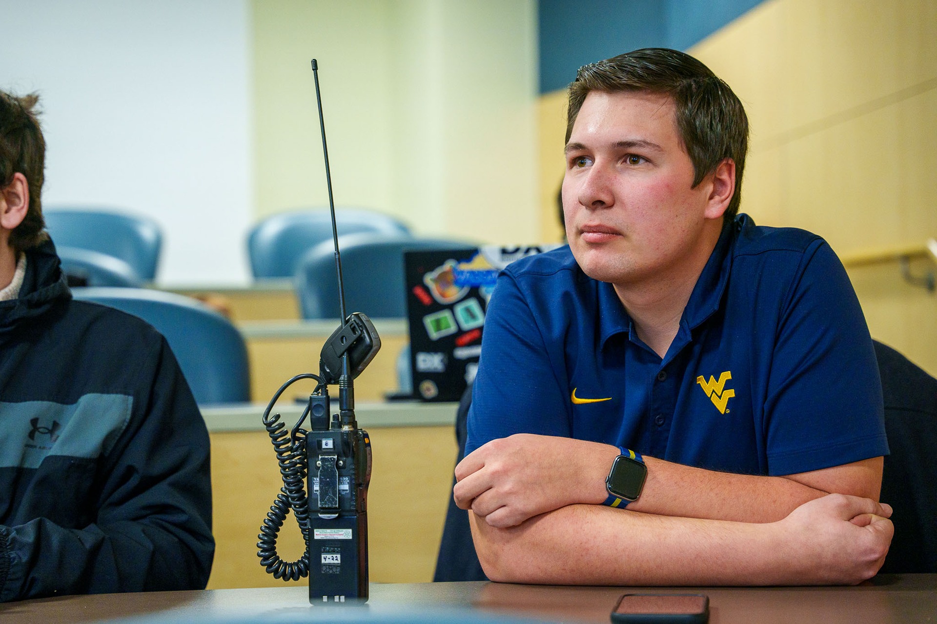 A student wearing a wvu polo sits in a classroom for a AM Radio Club meeting with a radio device on the table.
